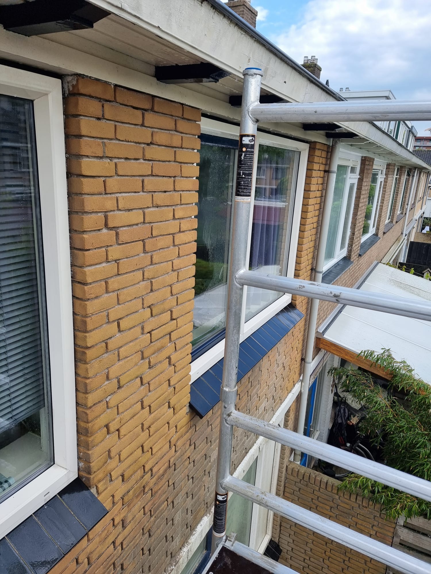 Steigerwerk langs gele bakstenen gevel met hemelwaterafvoer en kozijnen Steigerwerk langs bakstenen gevel voor constructief gevelherstel rondom kozijnen
