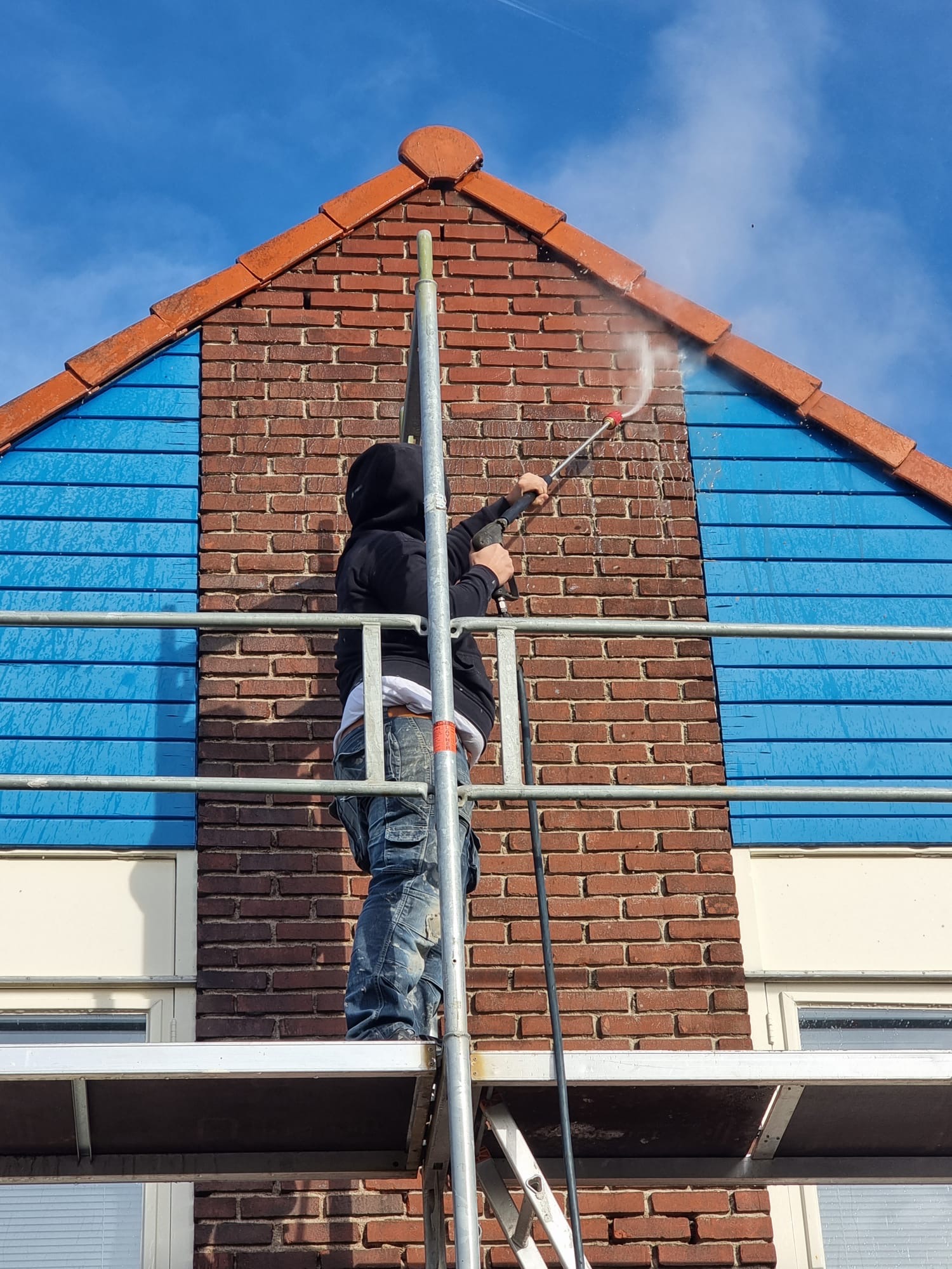 Vakman herstelt voegwerk aan rode bakstenen kopgevel met blauwe gevelbescherming Gevelreiniging combineren met voegwerk herstellen